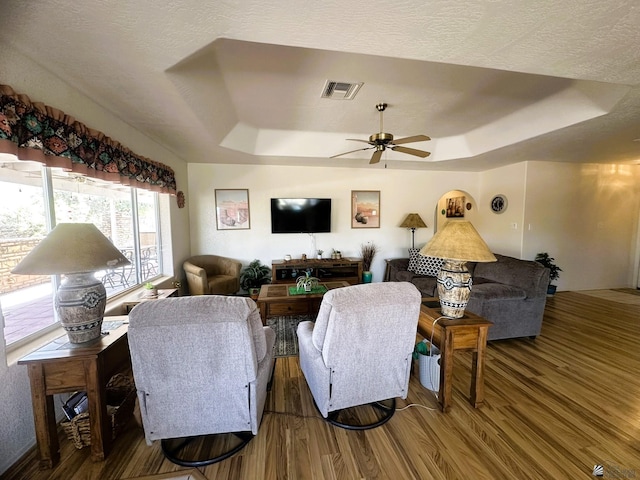 living room with a tray ceiling, wood finished floors, visible vents, and a ceiling fan