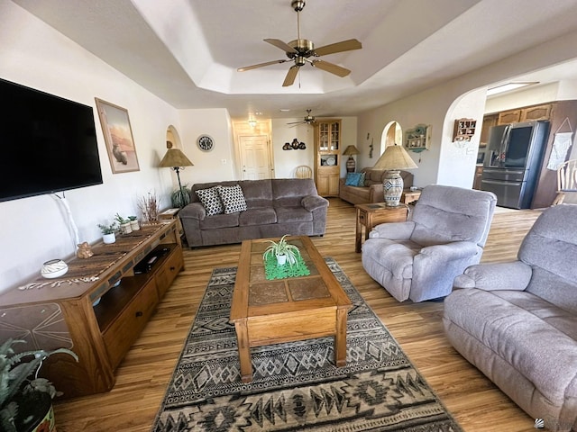 living room with light wood finished floors, ceiling fan, a tray ceiling, and arched walkways