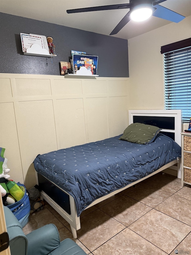 tiled bedroom featuring ceiling fan, a decorative wall, and wainscoting