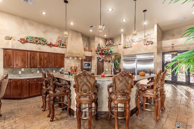 kitchen with a breakfast bar area, hanging light fixtures, built in appliances, light stone countertops, and premium range hood