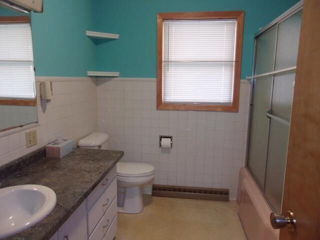 full bathroom with combined bath / shower with glass door, large vanity, toilet, and tile walls