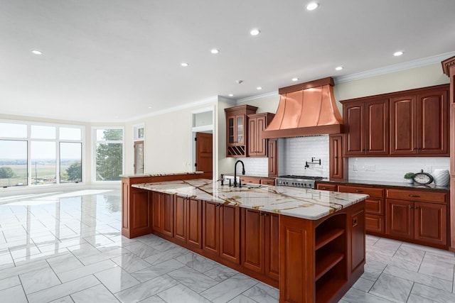 kitchen featuring tasteful backsplash, light stone counters, an island with sink, premium range hood, and sink