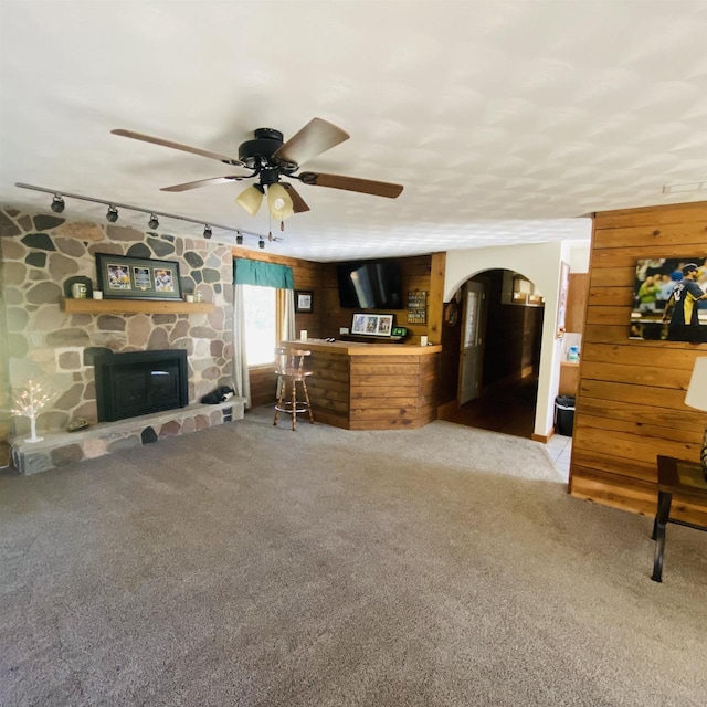 unfurnished living room featuring wood walls, a stone fireplace, ceiling fan, rail lighting, and carpet