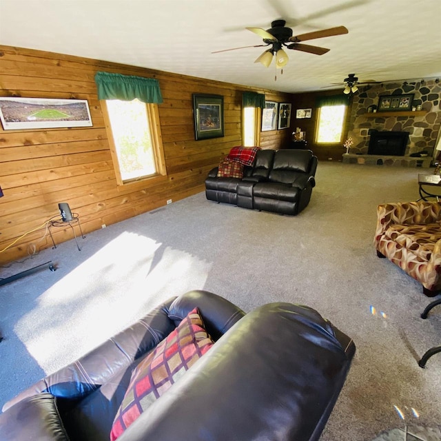 carpeted living room with a healthy amount of sunlight, ceiling fan, a stone fireplace, and wooden walls