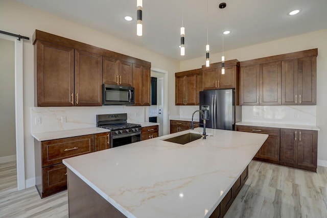 kitchen with an island with sink, sink, light stone counters, hanging light fixtures, and stainless steel appliances