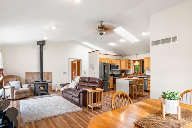 living room featuring a wood stove, ceiling fan, light hardwood / wood-style floors, a skylight, and high vaulted ceiling