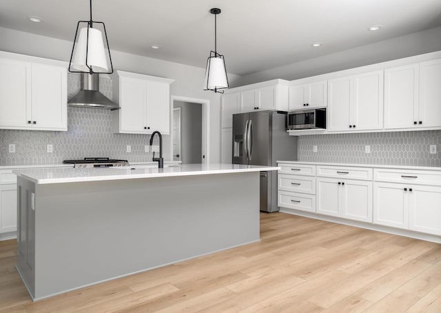 kitchen featuring a kitchen island with sink, stainless steel appliances, wall chimney range hood, and decorative light fixtures