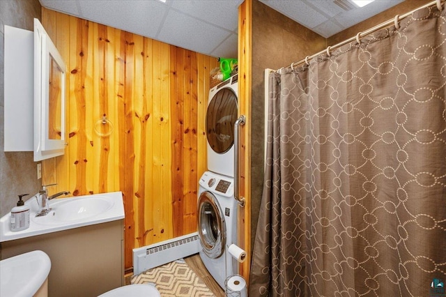 bathroom with toilet, a drop ceiling, wood walls, stacked washer and dryer, and baseboard heating