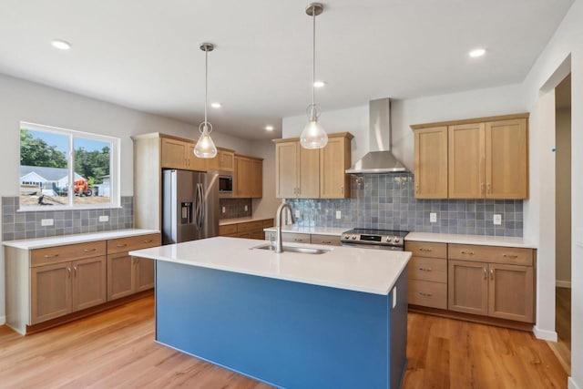 kitchen with sink, hanging light fixtures, appliances with stainless steel finishes, a kitchen island with sink, and wall chimney range hood