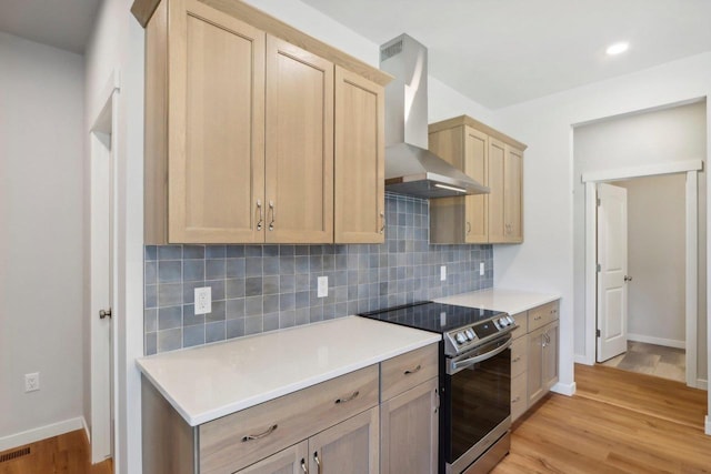 kitchen featuring tasteful backsplash, stainless steel range with electric cooktop, wall chimney range hood, light brown cabinets, and light wood-type flooring