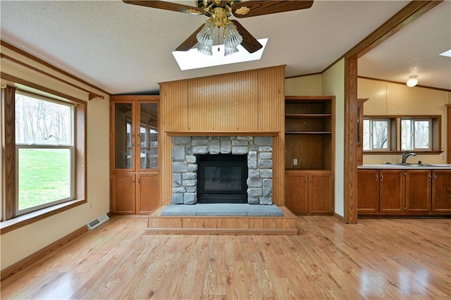 unfurnished living room featuring a stone fireplace, ceiling fan, vaulted ceiling with skylight, and light wood-type flooring