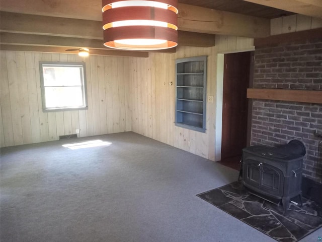 unfurnished living room with beamed ceiling, dark colored carpet, wooden walls, and a wood stove