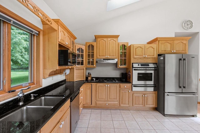 kitchen with sink, vaulted ceiling with skylight, dark stone countertops, light tile patterned floors, and appliances with stainless steel finishes