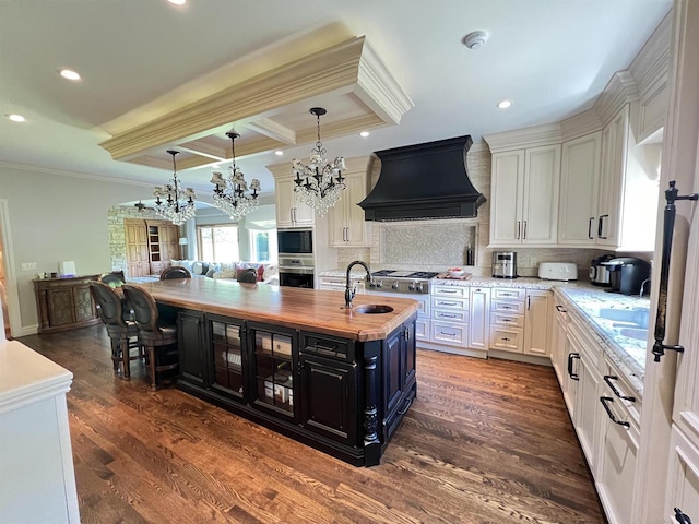 kitchen featuring premium range hood, dark wood-type flooring, a center island with sink, a notable chandelier, and butcher block countertops