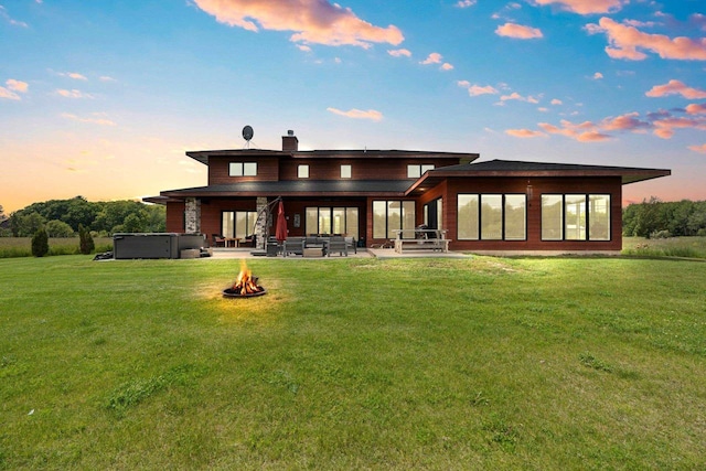 back house at dusk featuring a hot tub, a fire pit, a patio area, and a lawn