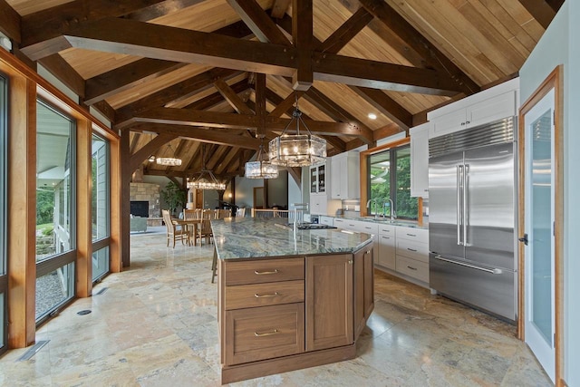 kitchen with dark stone counters, built in refrigerator, a large island, beam ceiling, and white cabinetry