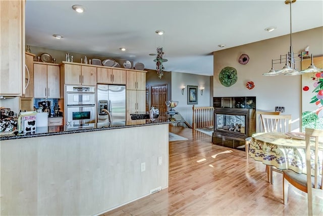 kitchen with light brown cabinetry, dark stone countertops, stainless steel appliances, a peninsula, and light wood finished floors