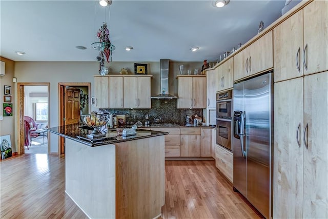 kitchen featuring light brown cabinetry, dark stone counters, wall chimney exhaust hood, stainless steel appliances, and a kitchen island