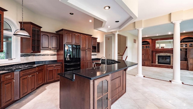 kitchen with decorative columns, stainless steel gas cooktop, an island with sink, black fridge, and dark stone counters