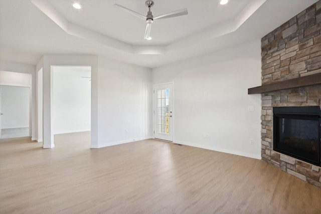 unfurnished living room featuring a tray ceiling, light hardwood / wood-style flooring, ceiling fan, and a stone fireplace