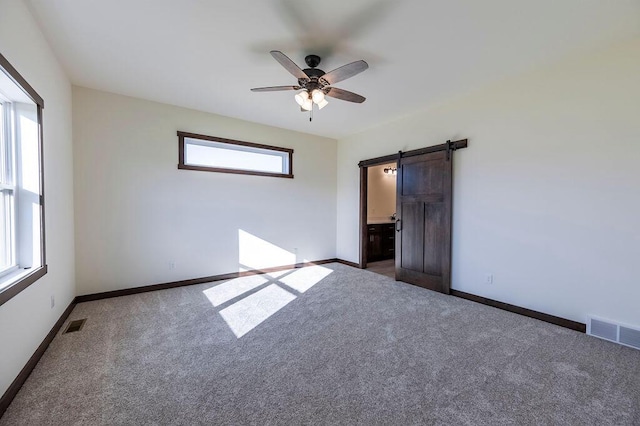 unfurnished bedroom featuring a barn door, dark carpet, ceiling fan, and ensuite bath