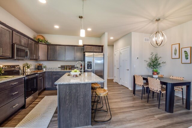 kitchen with light stone countertops, stainless steel appliances, hanging light fixtures, a kitchen island with sink, and dark brown cabinets