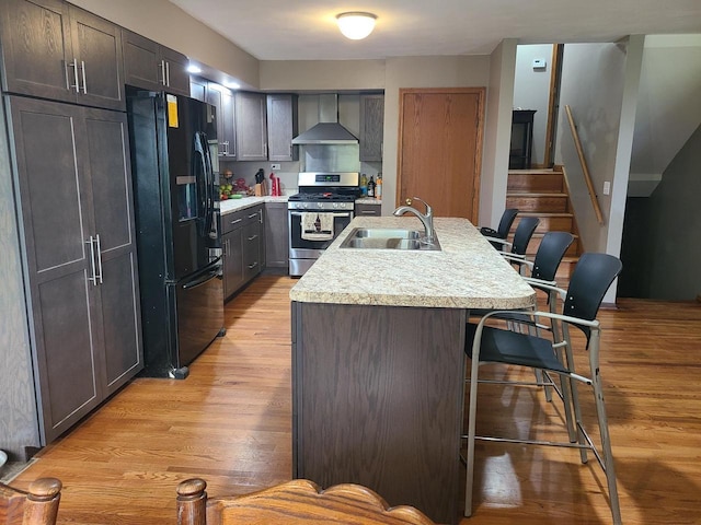 kitchen featuring a kitchen island with sink, a sink, wall chimney range hood, black refrigerator with ice dispenser, and gas stove