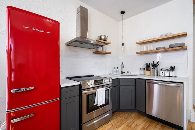 kitchen with appliances with stainless steel finishes, sink, hanging light fixtures, wall chimney range hood, and light wood-type flooring