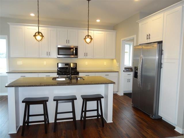 kitchen featuring pendant lighting, appliances with stainless steel finishes, a kitchen island with sink, dark stone countertops, and white cabinets