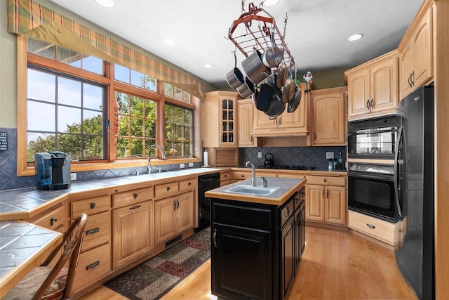 kitchen with sink, light hardwood / wood-style flooring, tile counters, a kitchen island with sink, and black appliances