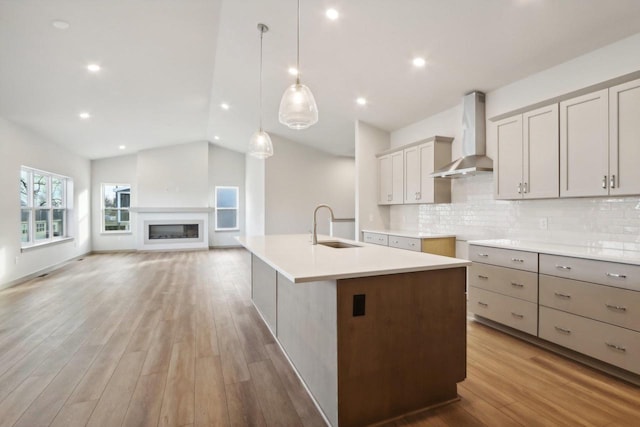 kitchen featuring sink, light wood-type flooring, wall chimney exhaust hood, hanging light fixtures, and an island with sink