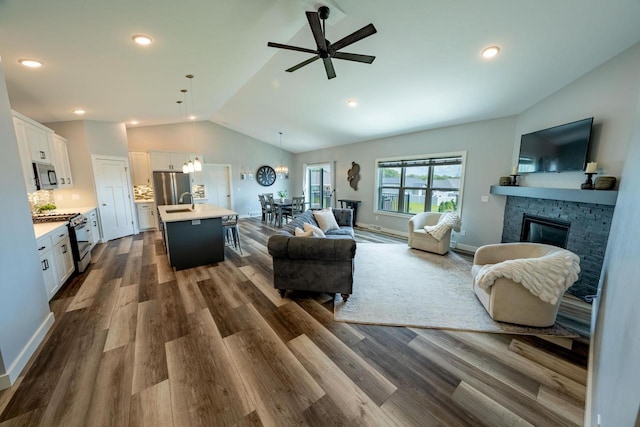 living room featuring dark wood-type flooring, vaulted ceiling, a stone fireplace, and ceiling fan with notable chandelier