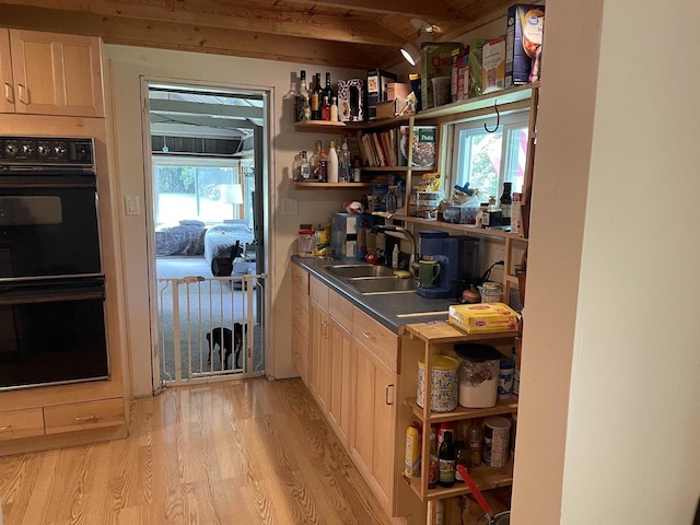 kitchen featuring black double oven, sink, light wood-type flooring, vaulted ceiling with beams, and light brown cabinetry