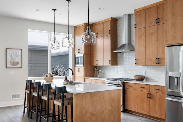 kitchen with stainless steel appliances, an island with sink, wall chimney exhaust hood, and light wood-type flooring