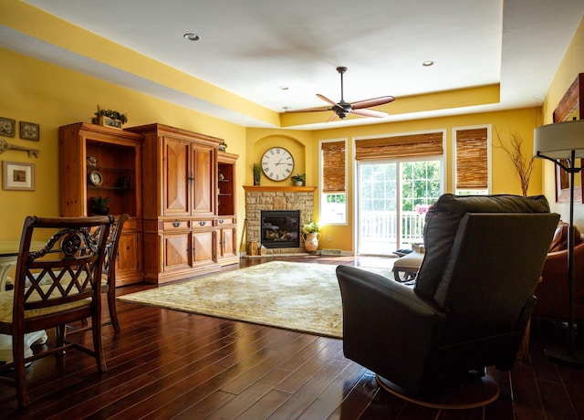 living area featuring a ceiling fan, a fireplace, a tray ceiling, and dark wood-type flooring