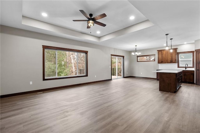 unfurnished living room featuring recessed lighting, ceiling fan with notable chandelier, baseboards, a tray ceiling, and light wood finished floors