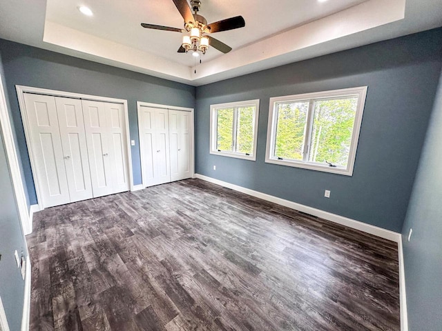 unfurnished bedroom with two closets, dark hardwood / wood-style flooring, ceiling fan, and a tray ceiling