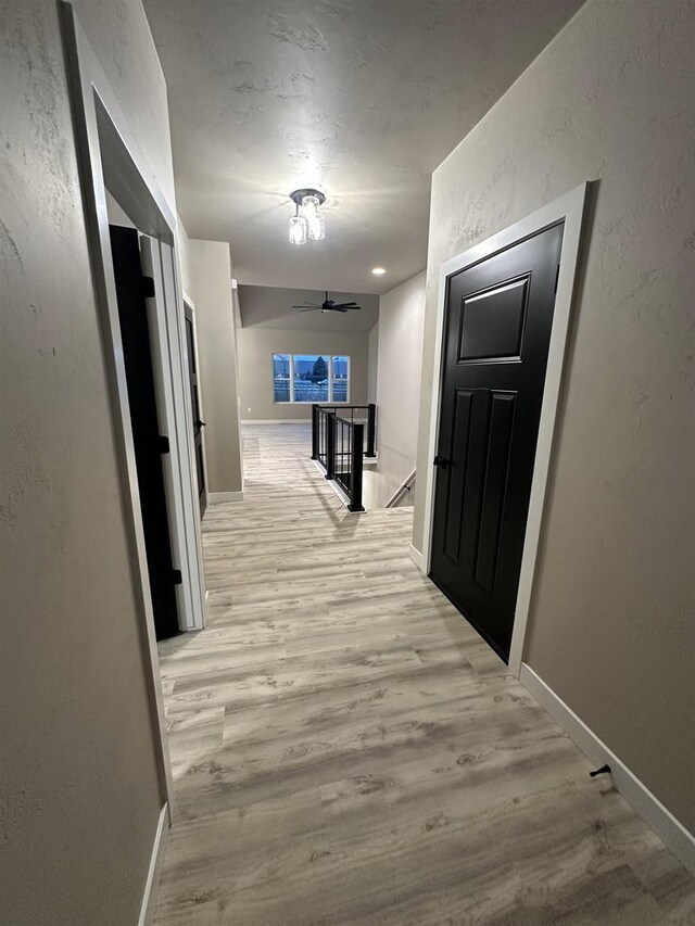 hall featuring light wood finished floors, a textured wall, baseboards, and an upstairs landing