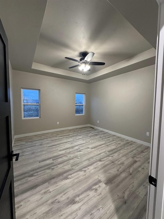 empty room with wood finished floors, a raised ceiling, a ceiling fan, and baseboards