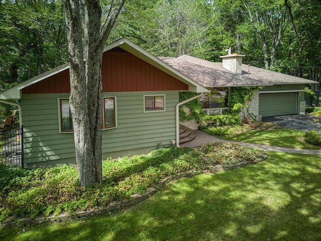 view of side of home with driveway, an attached garage, a shingled roof, a chimney, and a lawn