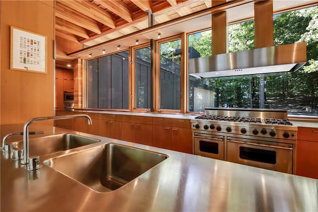 kitchen with range with two ovens, a sink, stainless steel countertops, wall chimney exhaust hood, and brown cabinets