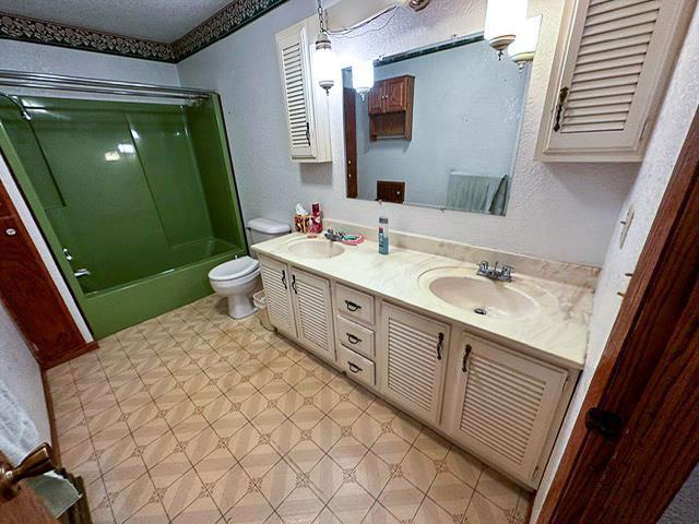 full bathroom with a sink, bath / shower combo with glass door, toilet, and double vanity