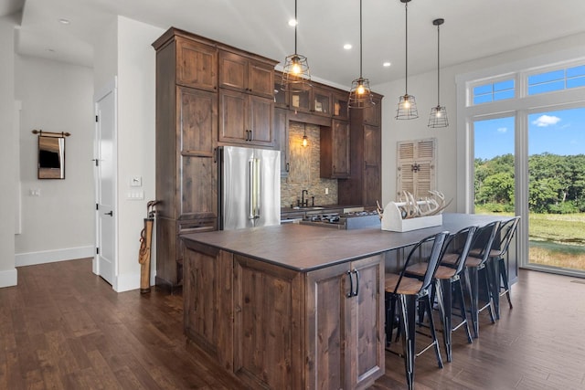 kitchen with high end fridge, dark brown cabinets, an island with sink, hanging light fixtures, and dark hardwood / wood-style floors