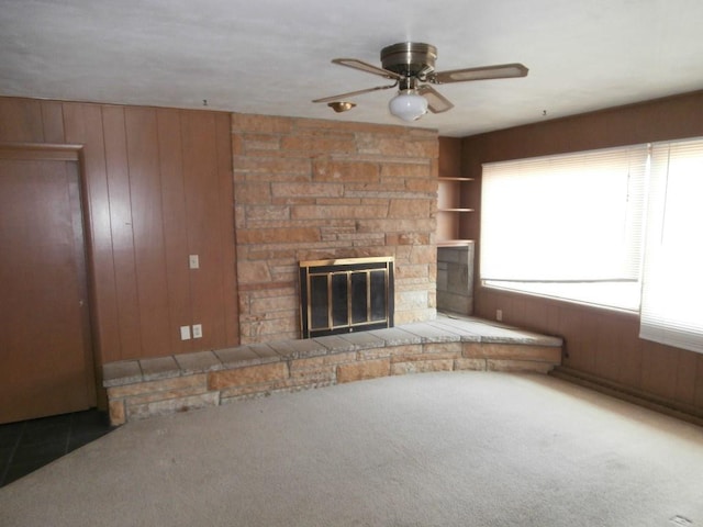 unfurnished living room with built in shelves, a stone fireplace, wood walls, carpet flooring, and ceiling fan