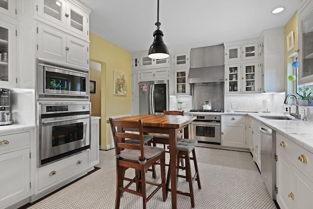 kitchen with white cabinetry, stainless steel appliances, decorative light fixtures, sink, and wall chimney range hood