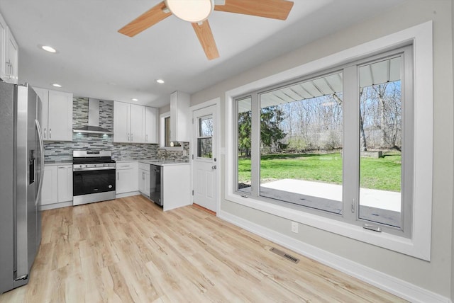 kitchen with wall chimney range hood, light hardwood / wood-style flooring, white cabinets, appliances with stainless steel finishes, and tasteful backsplash