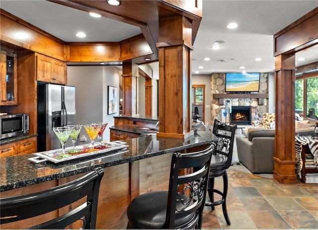 kitchen featuring appliances with stainless steel finishes, dark stone countertops, a fireplace, a kitchen bar, and decorative columns