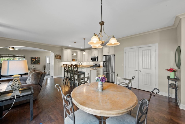 dining space with sink, ceiling fan with notable chandelier, crown molding, and dark wood-type flooring