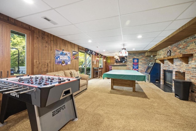 game room featuring brick wall, a brick fireplace, a drop ceiling, wooden walls, and light carpet