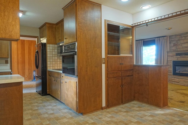 kitchen with a textured ceiling, black appliances, a stone fireplace, and decorative backsplash